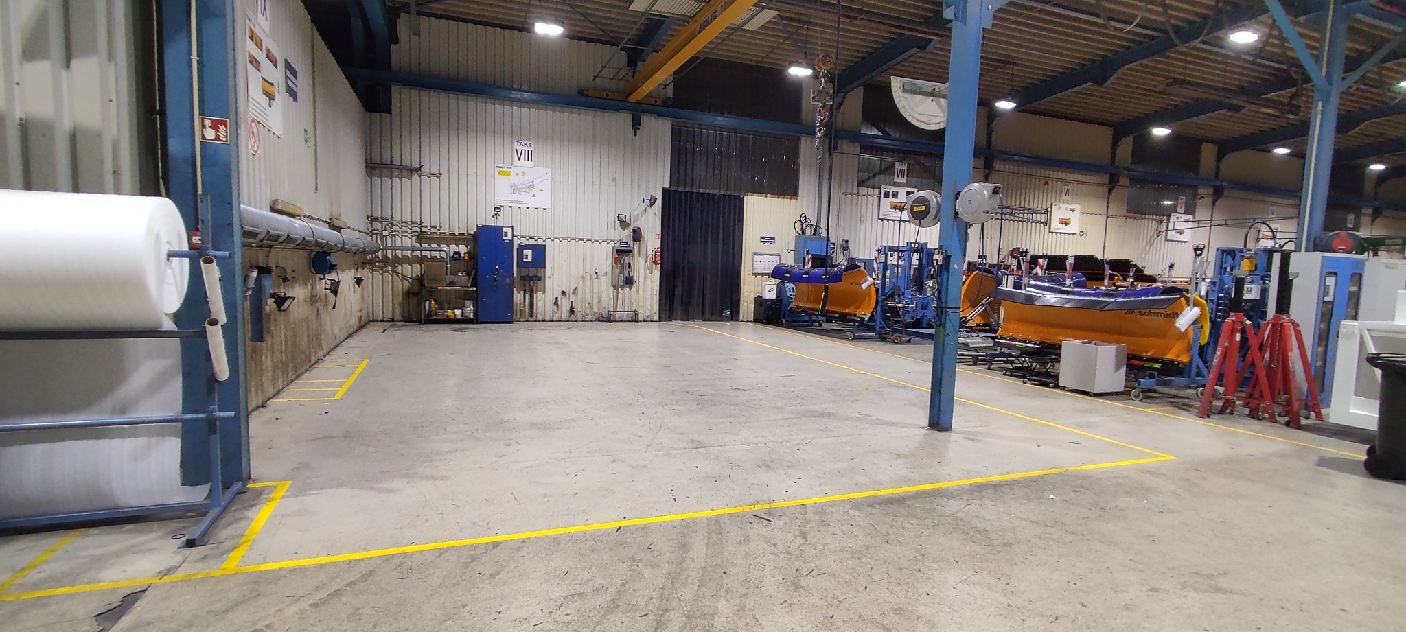 Wnętrze hali produkcyjnej z maszynami, rolką materiału i wyznaczonymi żółtymi liniami na podłodze.
