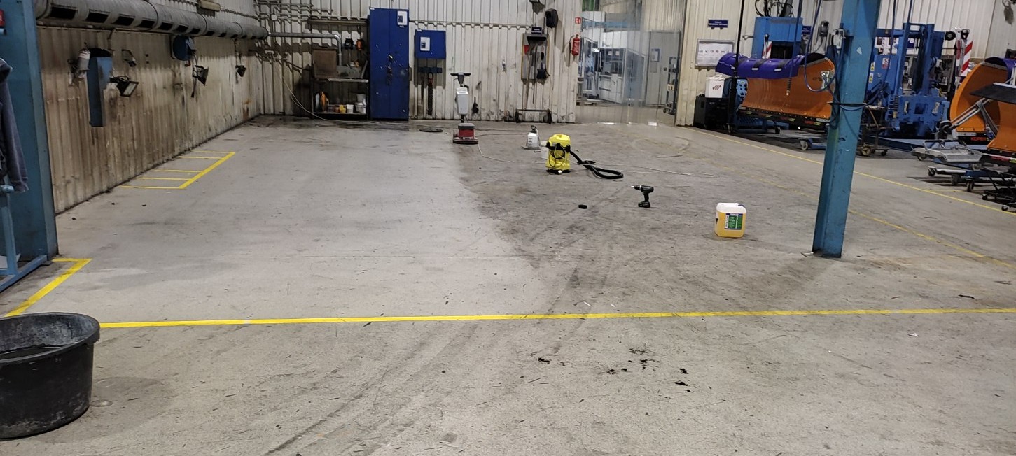 Betonowa posadzka hali przemysłowej częściowo umyta, z widocznym brudem, narzędziami (wiertarka, odkurzacz przemysłowy) i pojemnikiem z płynem czyszczącym, wyznaczona żółtymi liniami.
