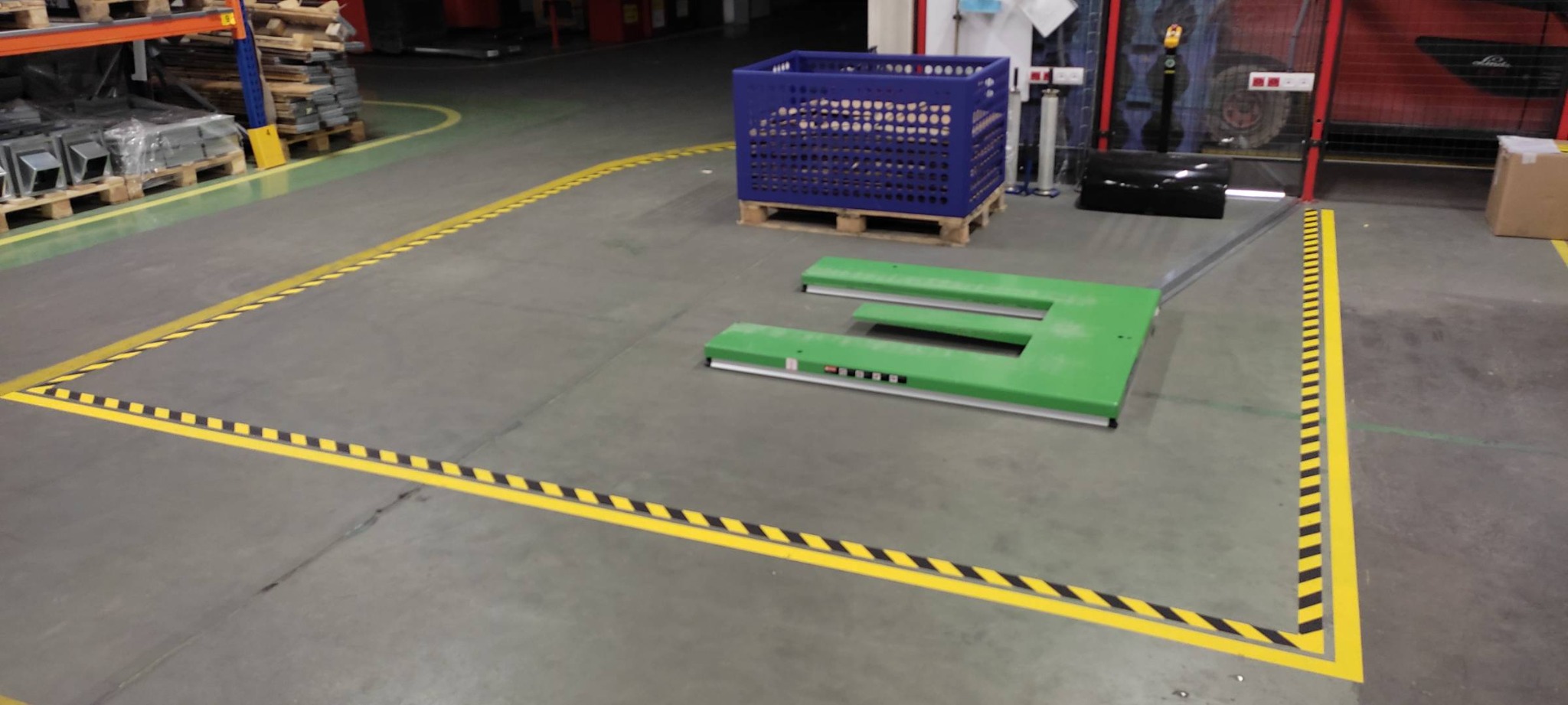 Oznakowane żółto-czarną taśmą stanowisko pracy w hali produkcyjnej, z zielonym podnośnikiem i niebieskim pojemnikiem na palecie w tle, widoczne regały magazynowe i wózek widłowy.
