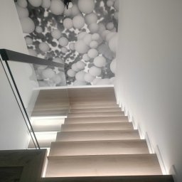 Dąbi - Nowoczesne schody z jasnego drewna z podświetleniem LED, szklaną balustradą i abstrakcyjną tapetą na suficie.