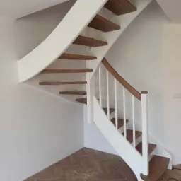 Nowoczesne schody z drewnianymi stopniami i białymi elementami konstrukcyjnymi, wkomponowane w jasne wnętrze z parkietem ułożonym we wzór jodełki.