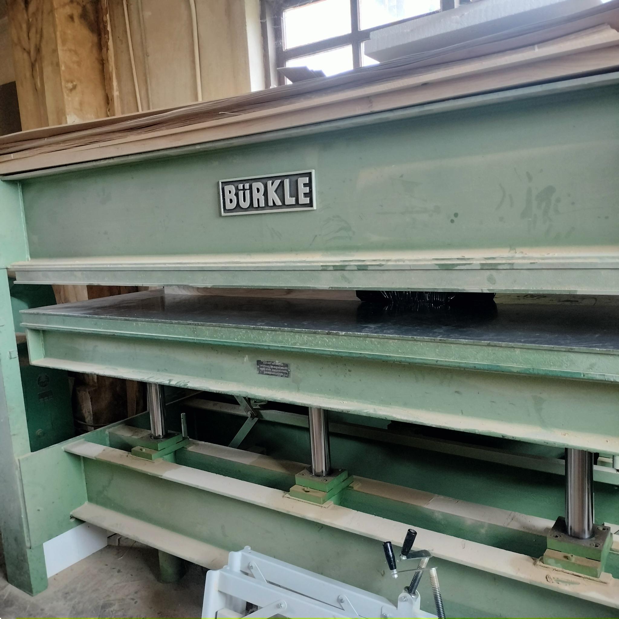 Hydrauliczna prasa do forniru marki Bürkle, z widocznymi warstwami materiału i elementami konstrukcyjnymi, w warsztacie.