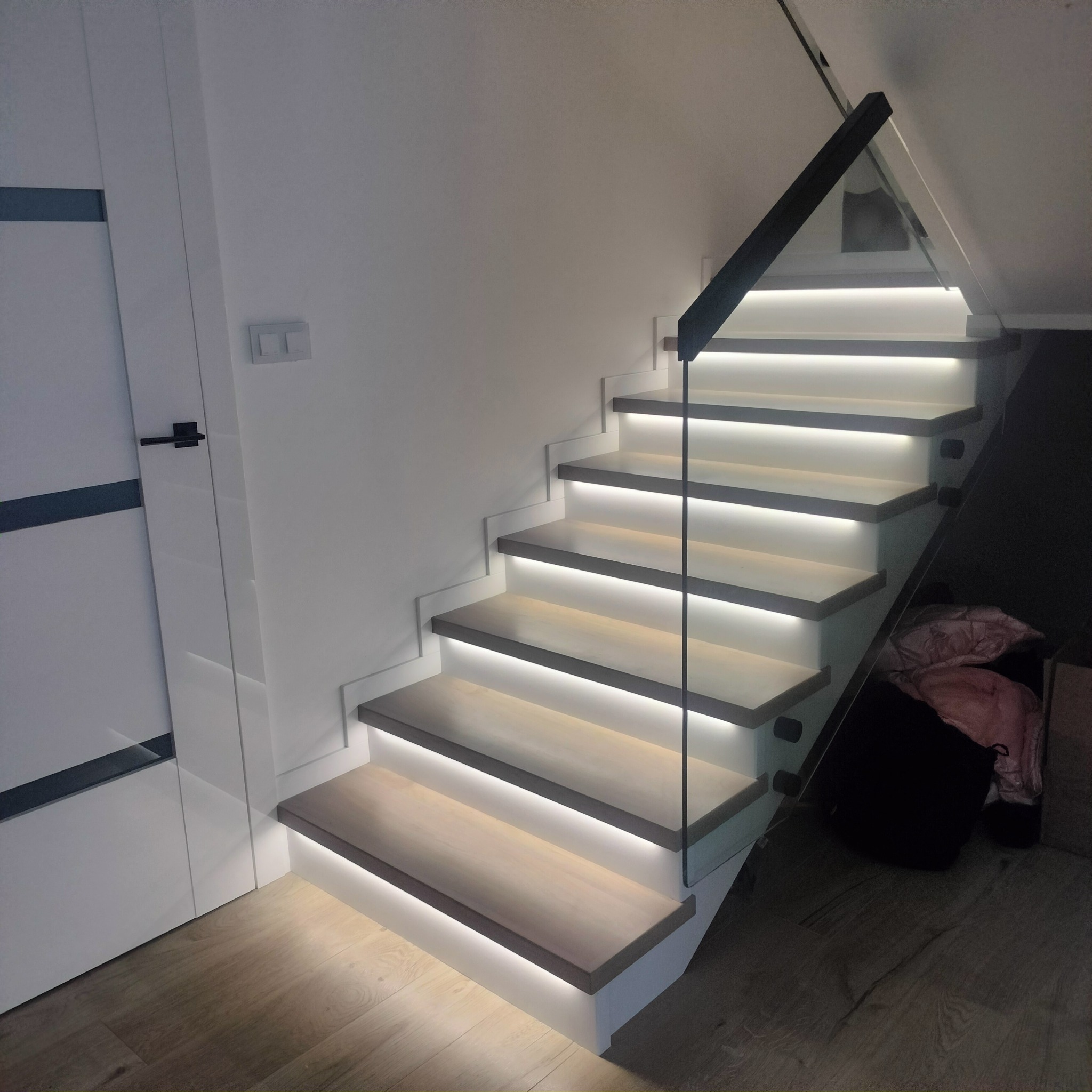 Nowoczesne schody drewniane z podświetleniem LED, szklaną balustradą i czarnymi elementami wykończeniowymi, widoczne z boku obok białych drzwi.