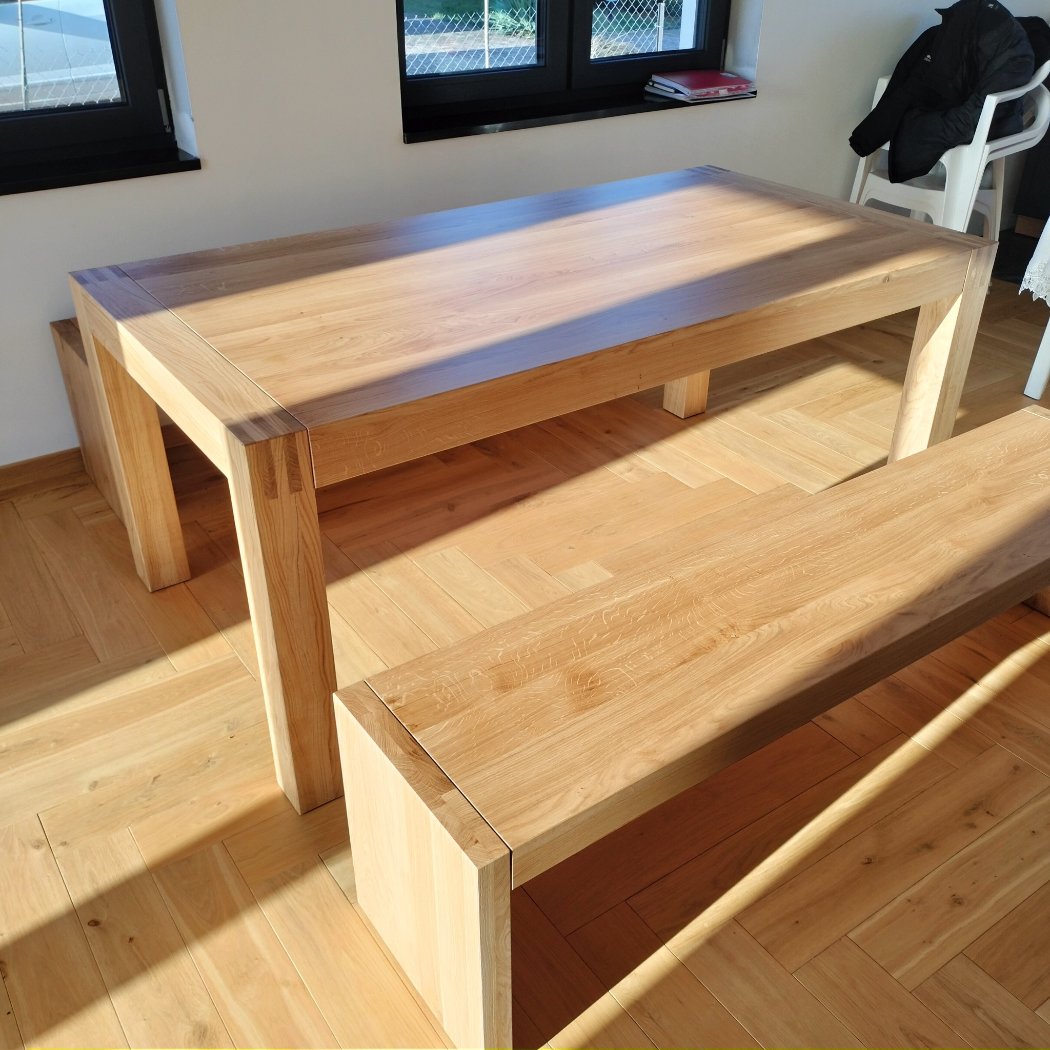 Jasny, dębowy stół z ławką w minimalistycznym stylu, ustawiony na parkiecie w jodełkę, oświetlony promieniami słońca wpadającymi przez okno.