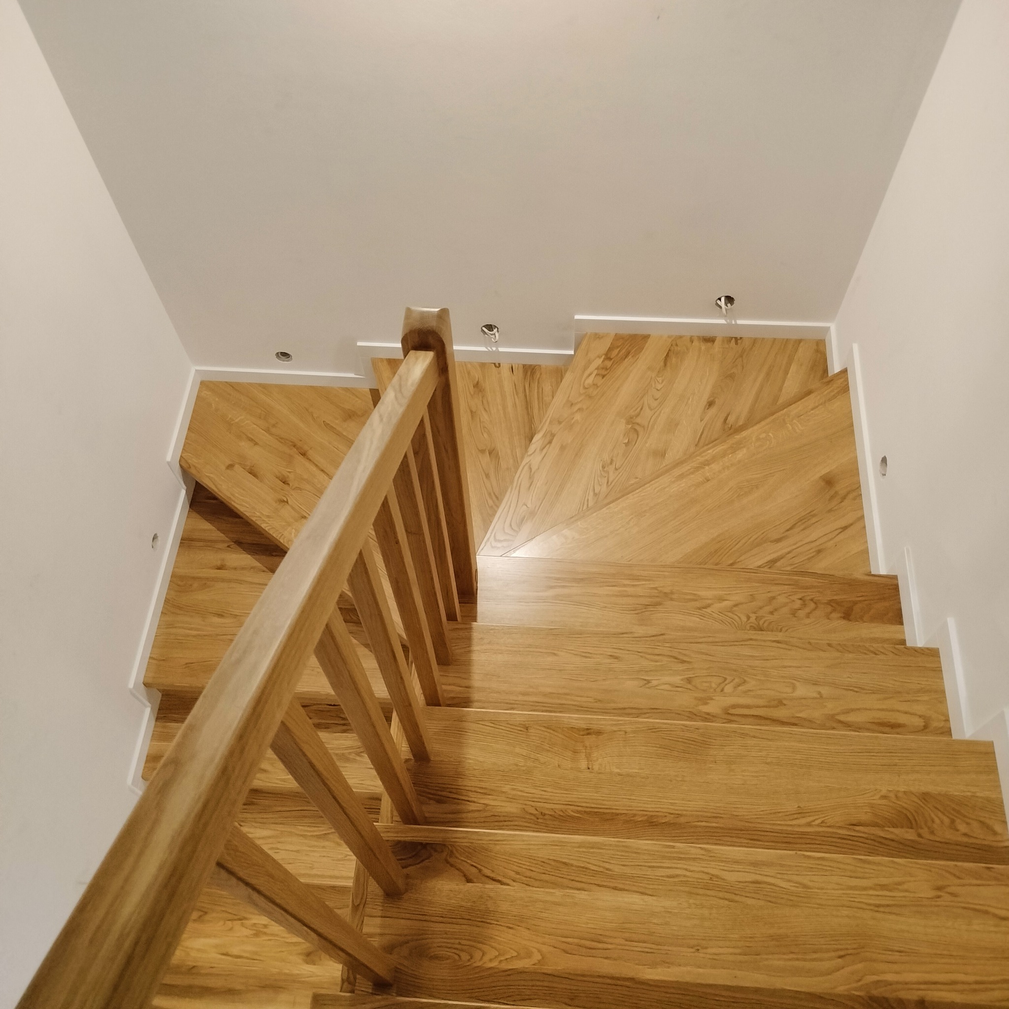 Widok z góry na jasne, drewniane schody z balustradą, w nowoczesnym wnętrzu z białymi ścianami i sufitami, z widocznymi punktami oświetleniowymi.