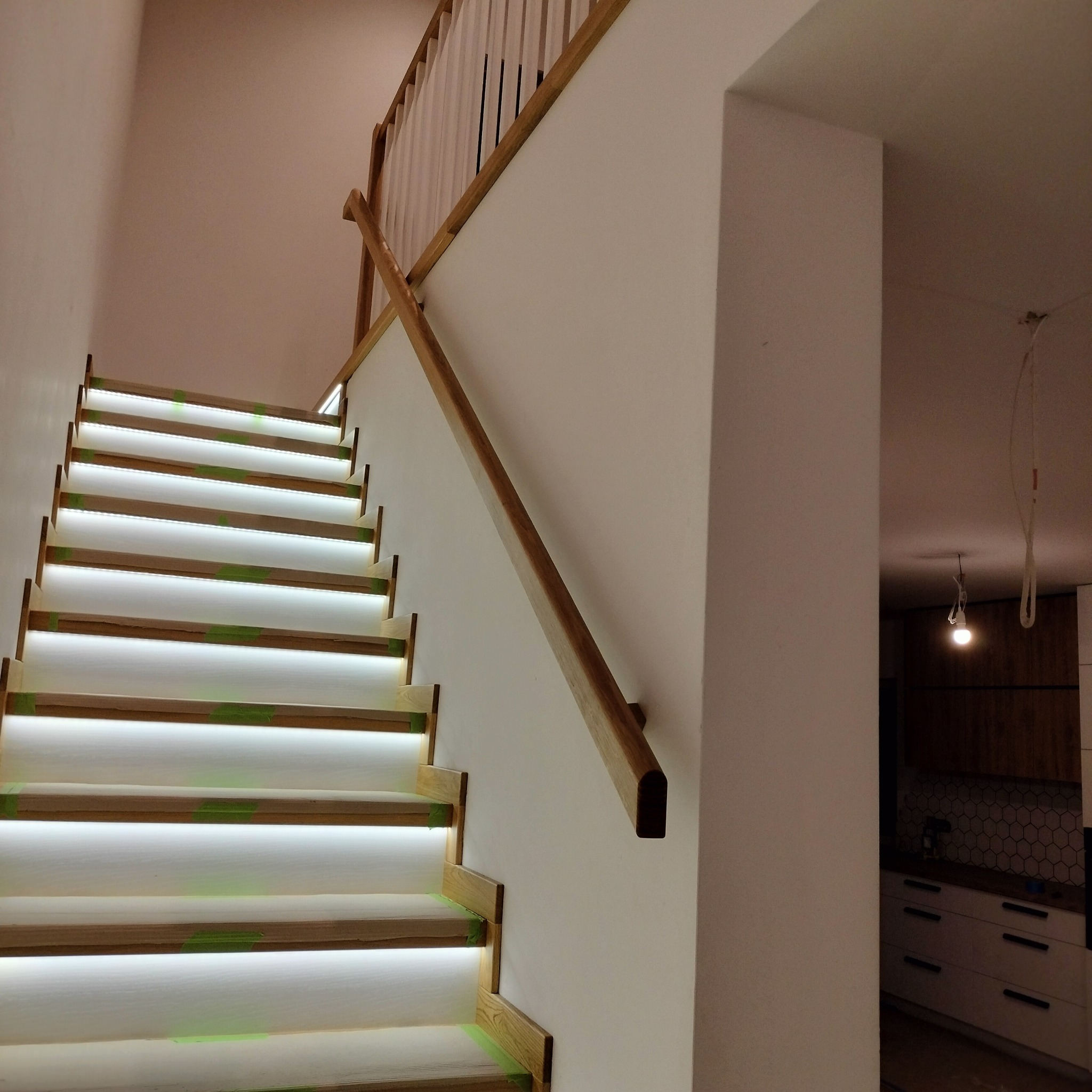 Nowoczesne schody z drewnianymi stopniami i podświetleniem LED, zabezpieczone taśmą malarską, widok z dołu na białą balustradę i fragment kuchni w tle.