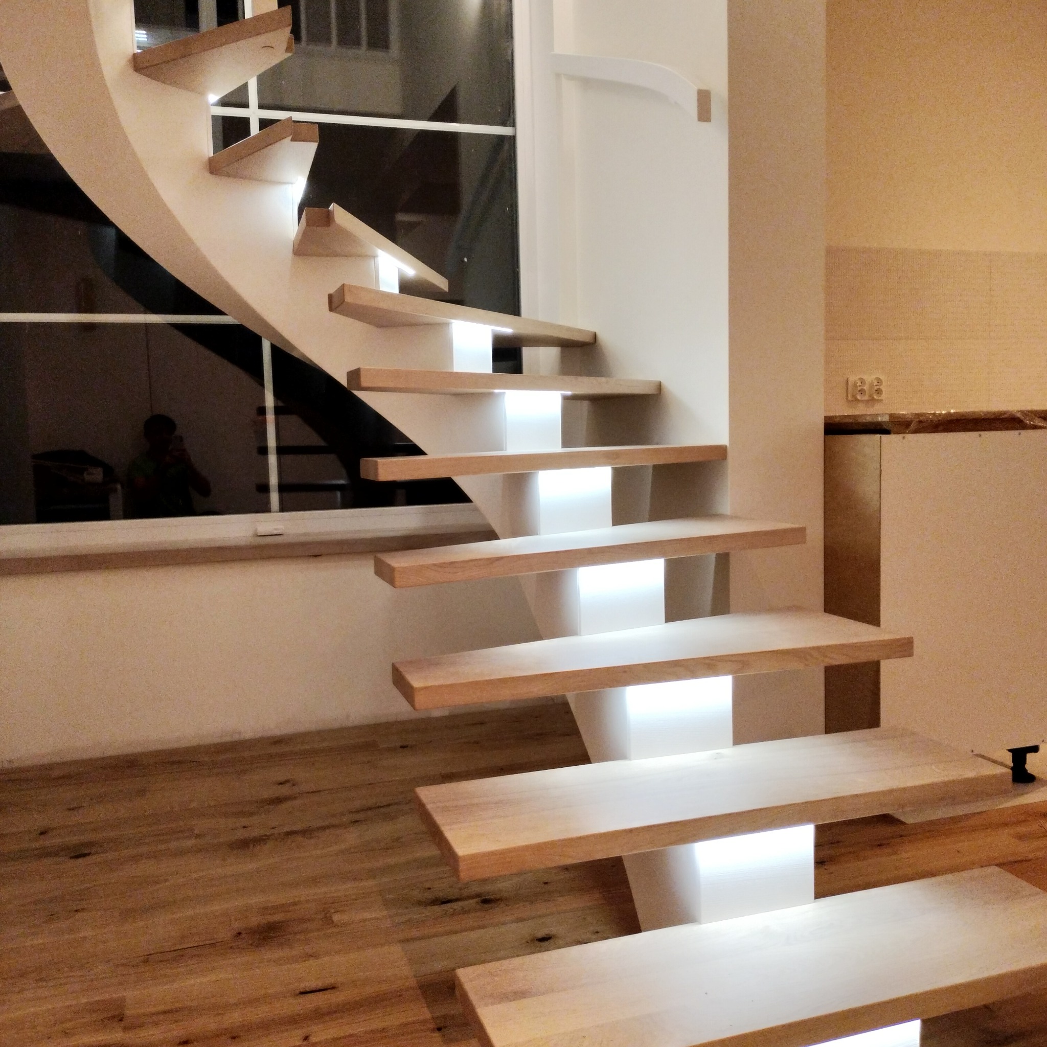 Nowoczesne, podświetlane stopnie schodów z jasnego drewna, wsporniki w kolorze białym, widoczne odbicie osoby w oknie.