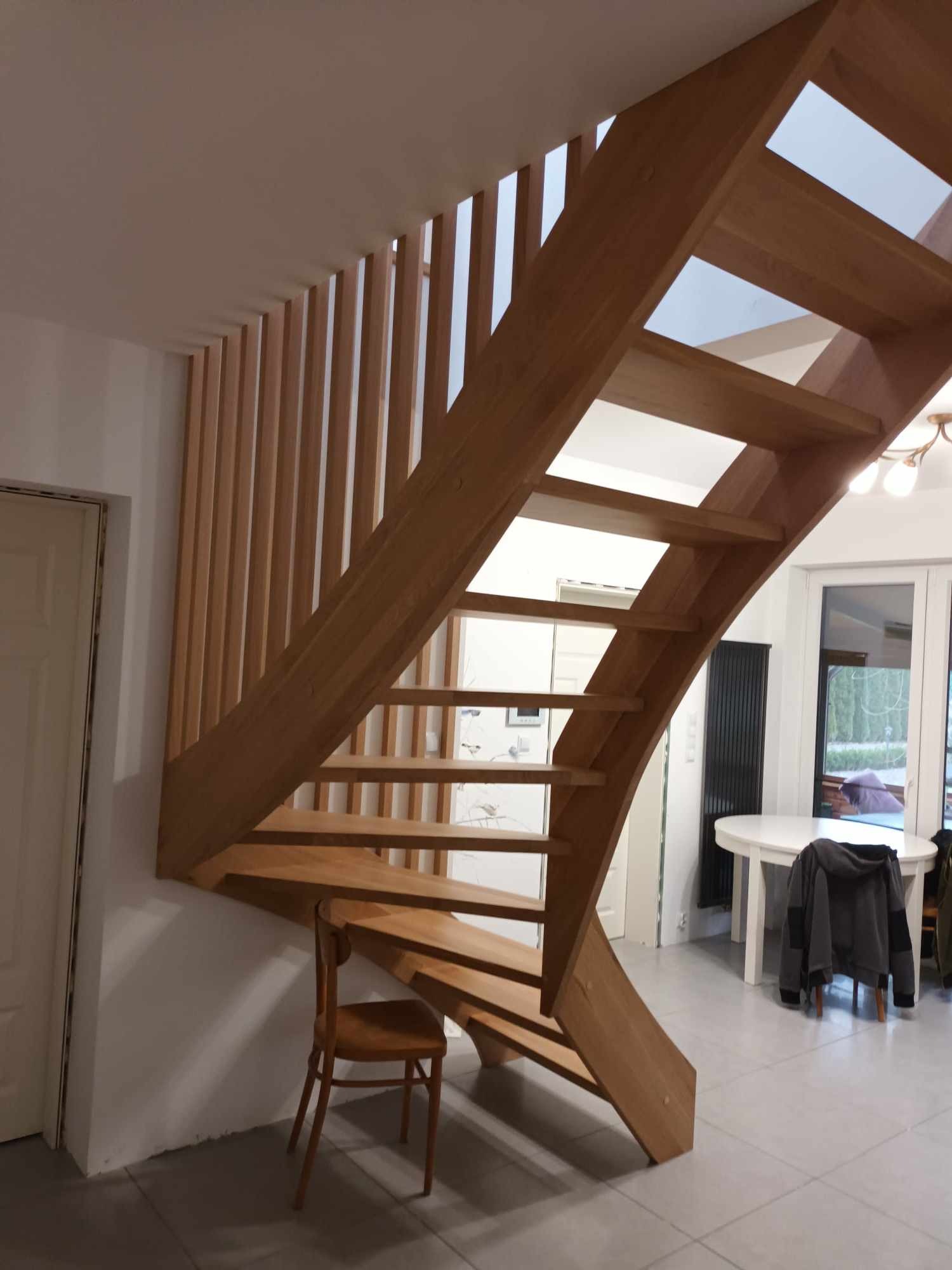 Drewniane schody z ażurową balustradą z pionowych listew, oświetlone światłem dziennym wpadającym przez okno, z krzesłem pod schodami.