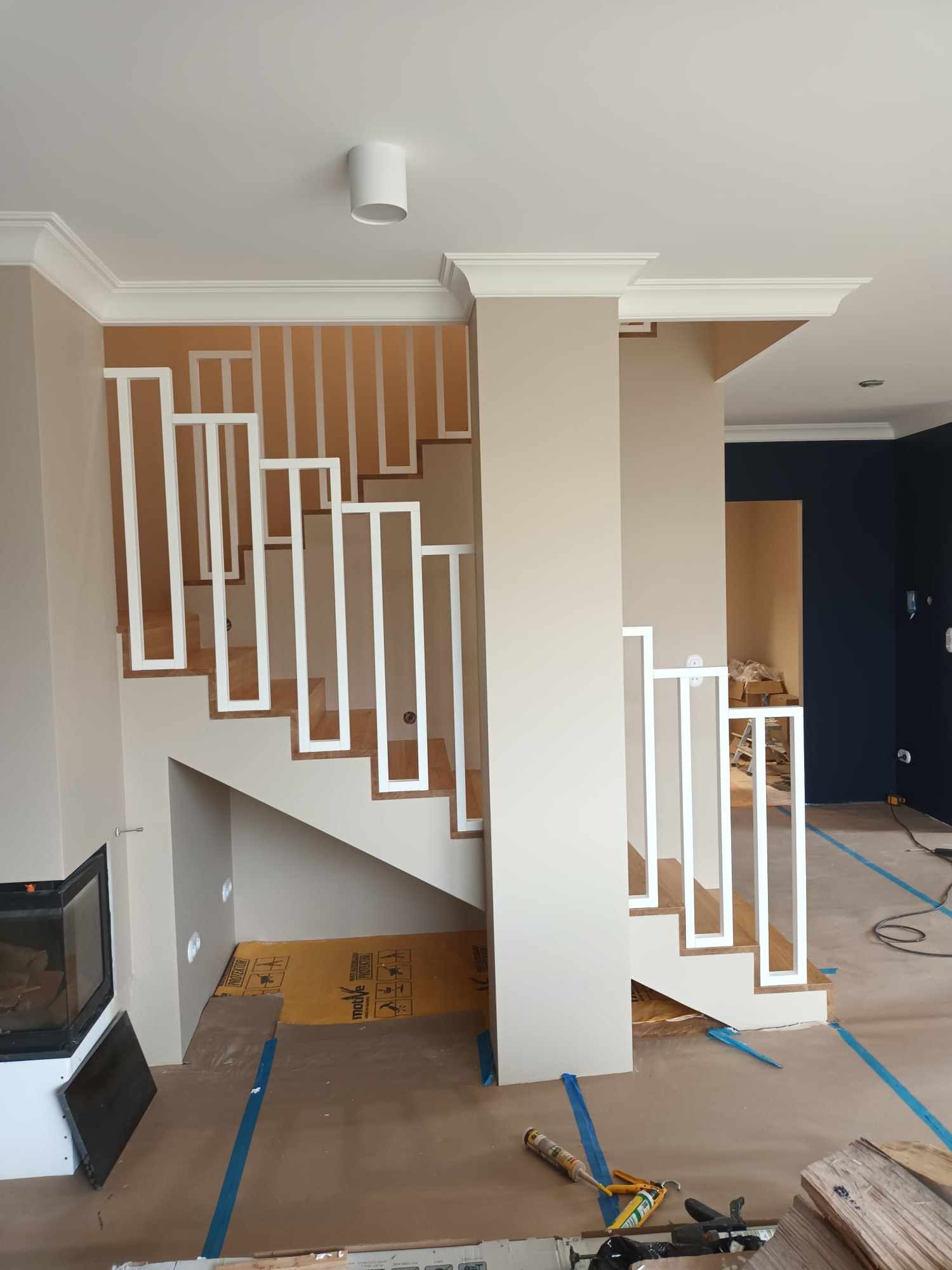 Nowoczesne schody z drewnianymi stopniami i białymi, geometrycznymi balustradami w trakcie montażu, widoczne narzędzia i zabezpieczenia podłogi.