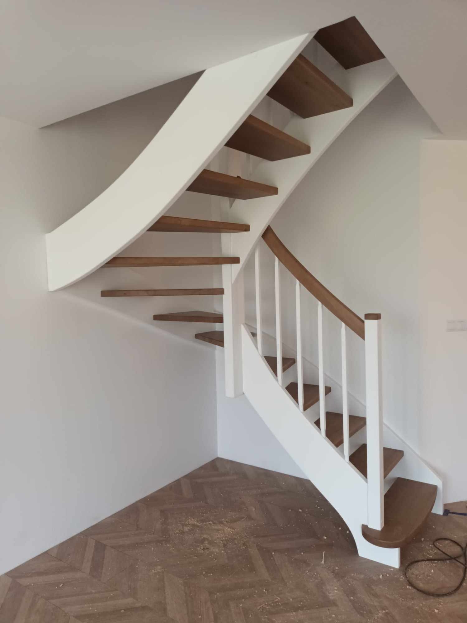 Nowoczesne schody z drewnianymi stopniami i białymi elementami konstrukcyjnymi, wkomponowane w jasne wnętrze z parkietem ułożonym we wzór jodełki.