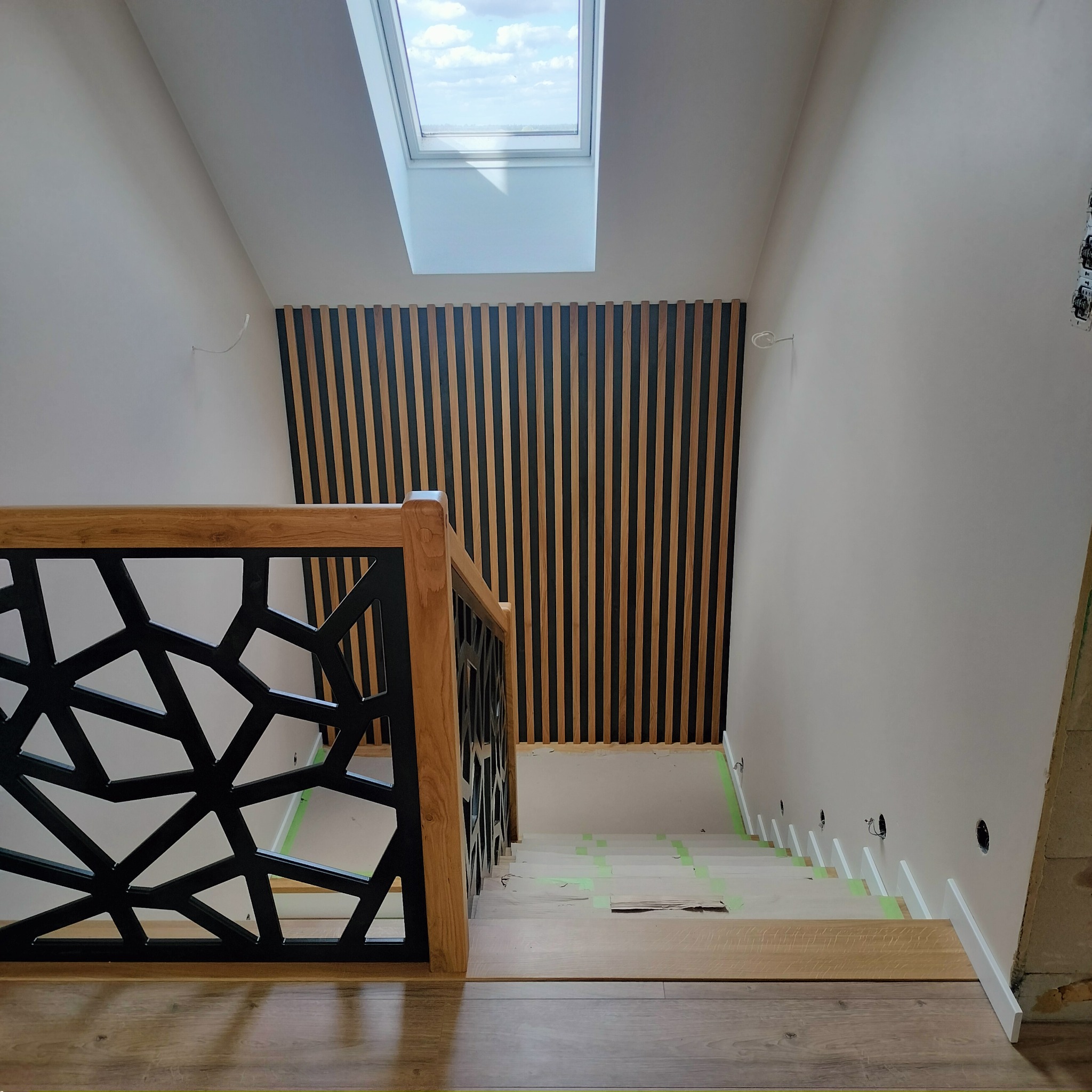 Wnętrze domu w trakcie wykańczania: widok z góry na drewniane schody z czarną, geometryczną balustradą, ściana z drewnianymi panelami pionowymi, okno dachowe i widoczne kable elektryczne.