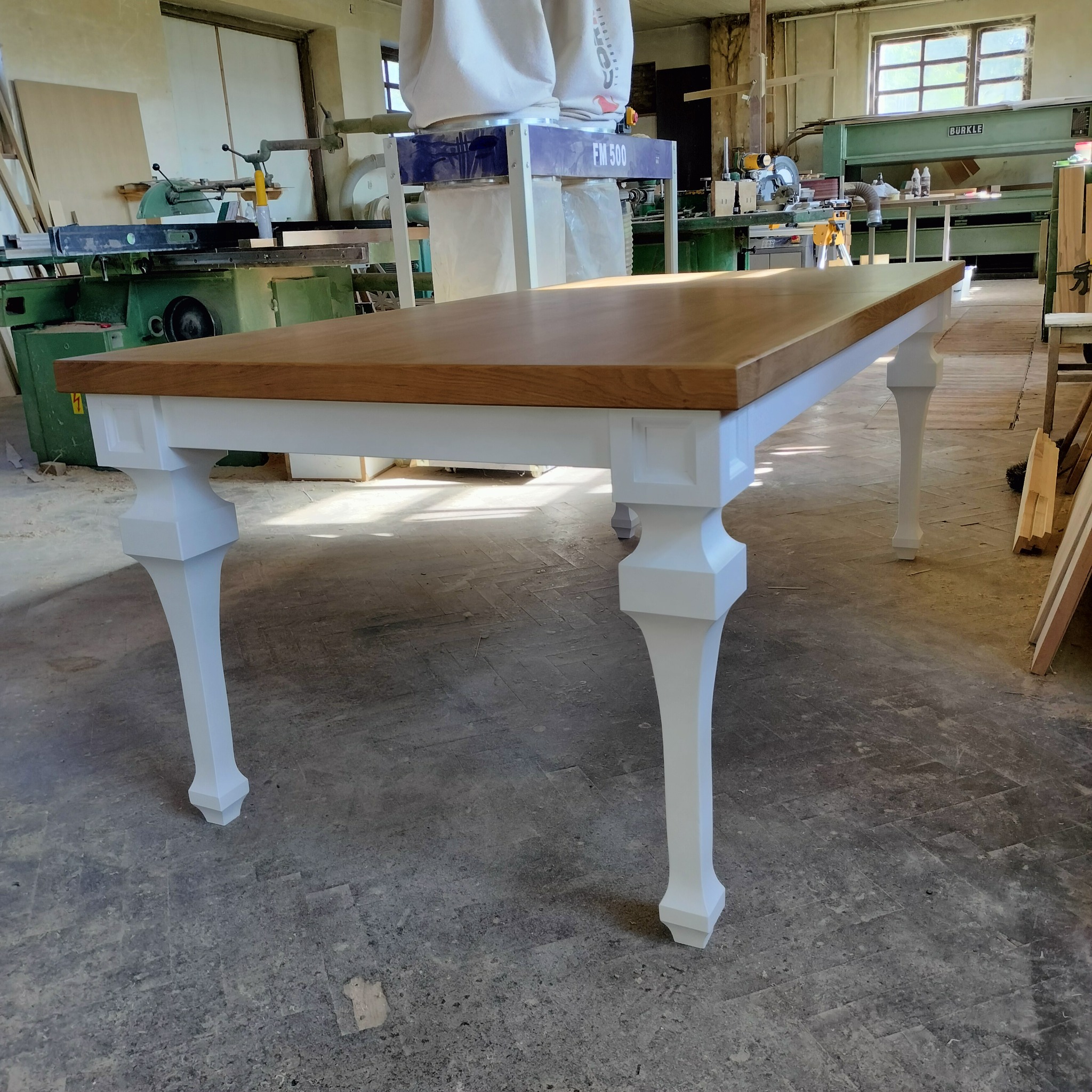 Stół z drewnianym blatem i białymi, toczonymi nogami, sfotografowany w warsztacie stolarskim na tle maszyn do obróbki drewna.