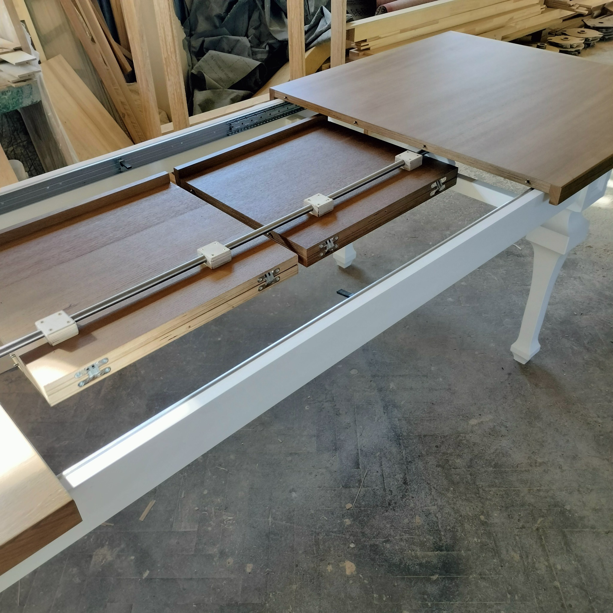 Rozsuwany stół z blatem w odcieniach brązu, mechanizm rozsuwania z metalowymi prowadnicami i białymi elementami mocującymi, widoczne drewniane elementy w tle.