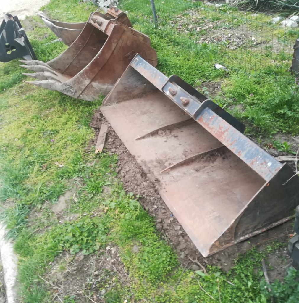 Rdzewiejące łyżki koparkowe o różnych rozmiarach i kształtach leżące na trawie, widoczne ślady użytkowania i ziemi.