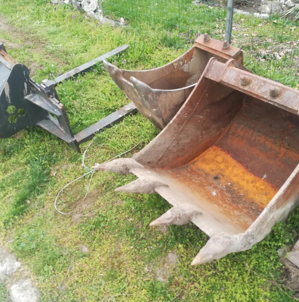 Dwa zardzewiałe, używane łyżki koparkowe z zębami i czarny osprzęt do koparki leżą na zielonej trawie.