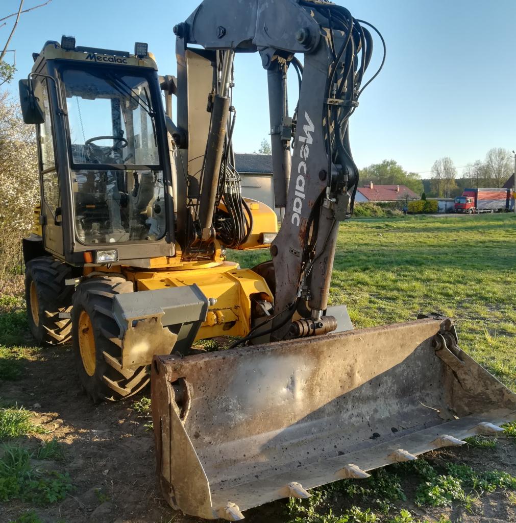 Żółto-szara koparka Mecalac z widocznymi śladami użytkowania na łyżce, stojąca na trawiastym terenie z budynkami w tle, oświetlona popołudniowym słońcem.