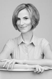 Portret kobiety w koszuli w paski, opierającej się o białe biurko, z delikatnym uśmiechem i krótkimi, falowanymi włosami w odcieniach szarości.