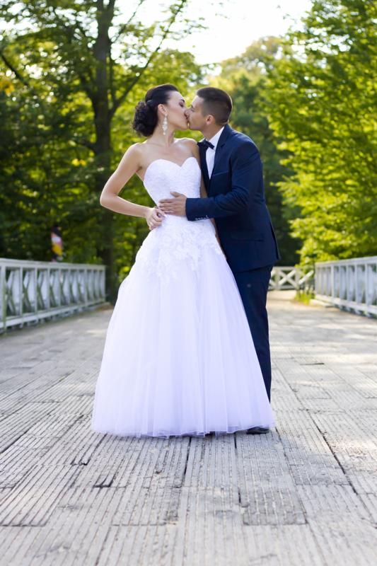 Para młoda całująca się na drewnianym moście otoczonym zielenią, panna młoda w białej sukni z koronkową górą i welonem, pan młody w granatowym garniturze z muchą.