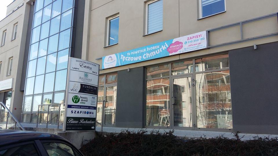 Fasada budynku z szyldem 'Tęczowe Chmurki', informującym o otwarciu placówki opiekuńczej; widoczne okna z odbiciami i tablica informacyjna z innymi firmami.