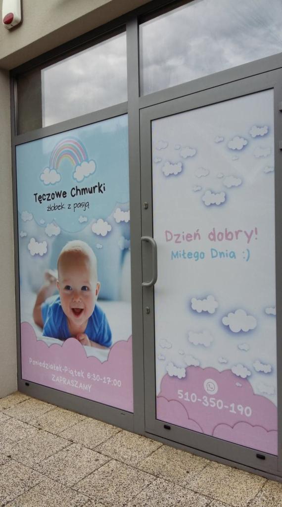 Wejście do placówki opiekuńczej z naklejonymi na szybach grafikami: uśmiechnięte niemowlę, tęcza, chmurki, godziny otwarcia i numer telefonu.