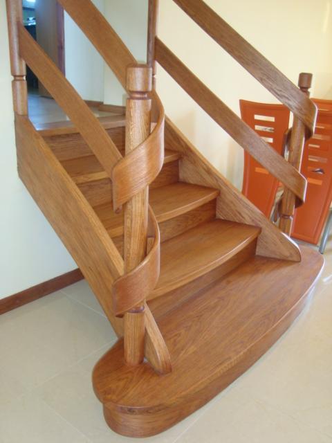 Drewniane schody z balustradą o nietypowym kształcie, oświetlone naturalnym światłem, z widoczną fakturą drewna i detalami wykończenia w tle jasne ściany i pomarańczowe krzesło.