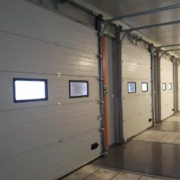 Wnętrze hali z rzędem białych segmentowych bram przemysłowych z małymi okienkami, widok z perspektywy podłogi.