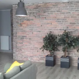 Wnętrze pokoju z odsłoniętą ceglaną ścianą, trzema roślinami w czarnych donicach, szarą sofą z żółtą poduszką oraz wiszącą lampą w stylu industrialnym.