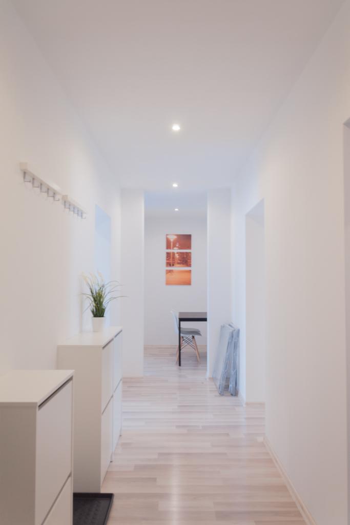 Jasny korytarz z białymi ścianami, drewnianą podłogą, białymi szafkami na buty i wieszakiem na ubrania, zakończony widokiem na pokój z obrazami na ścianie.