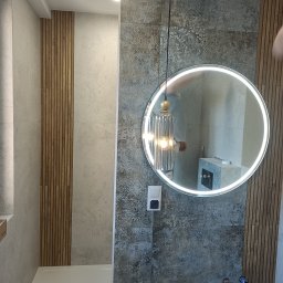 BUD-POL Krzysztof Chmiel - Nowoczesna łazienka z okrągłym lustrem LED, dekoracyjną lampą wiszącą, płytkami z wzorem i panelami drewnianymi na ścianie.