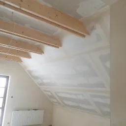 Poddasze w trakcie prac wykończeniowych, widoczne drewniane belki konstrukcyjne pod sufitem, częściowo pokryte płytami gipsowo-kartonowymi, szpachlowane łączenia, grzejnik pod oknem.
