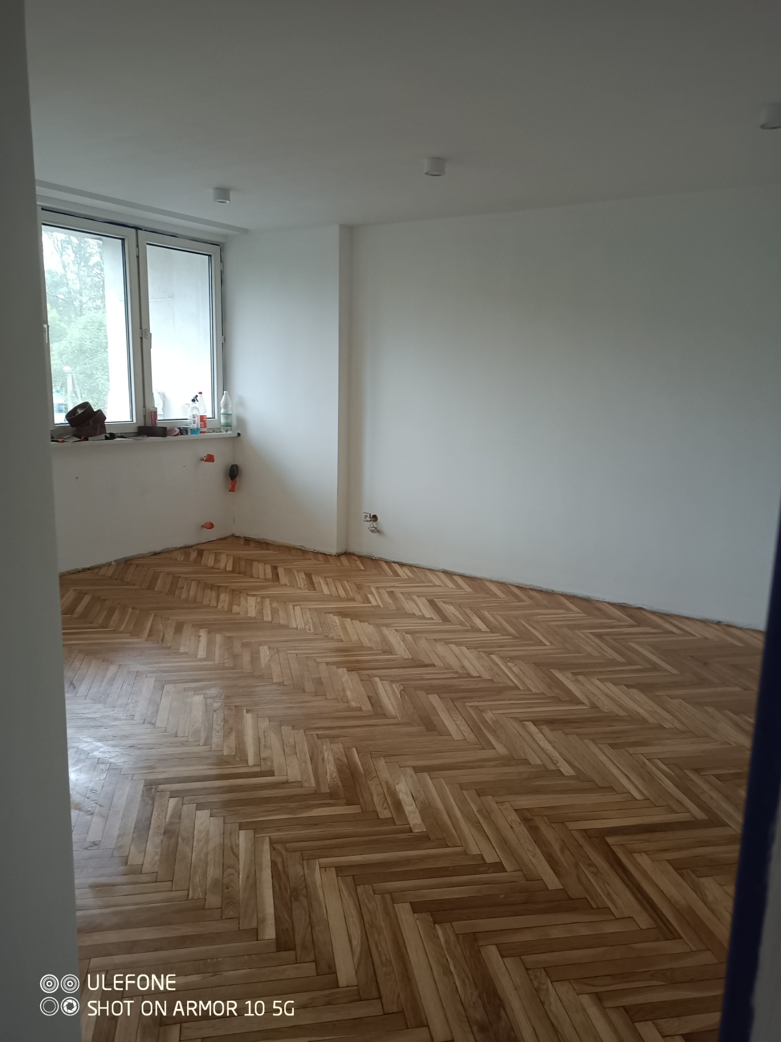 Jasne parkiety ułożone w jodełkę w pustym, odświeżonym pokoju z oknem. Widoczne ślady po remoncie przy ścianie i gniazdka elektryczne. Minimalistyczne wnętrze.