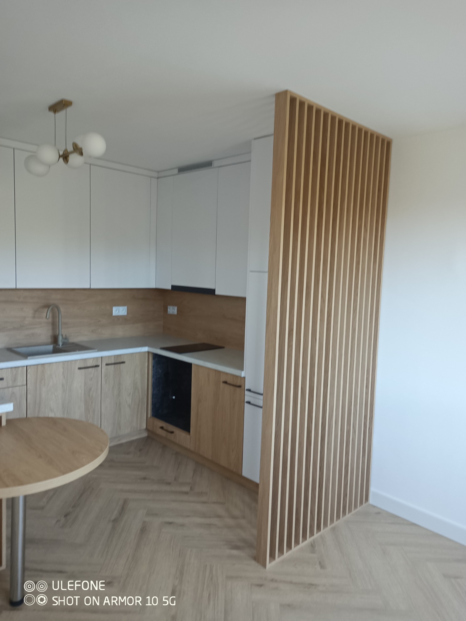 Jasna kuchnia z drewnianymi akcentami: zabudowa, podłoga w jodełkę i ażurowa ścianka działowa. Nowoczesne oświetlenie sufitowe.