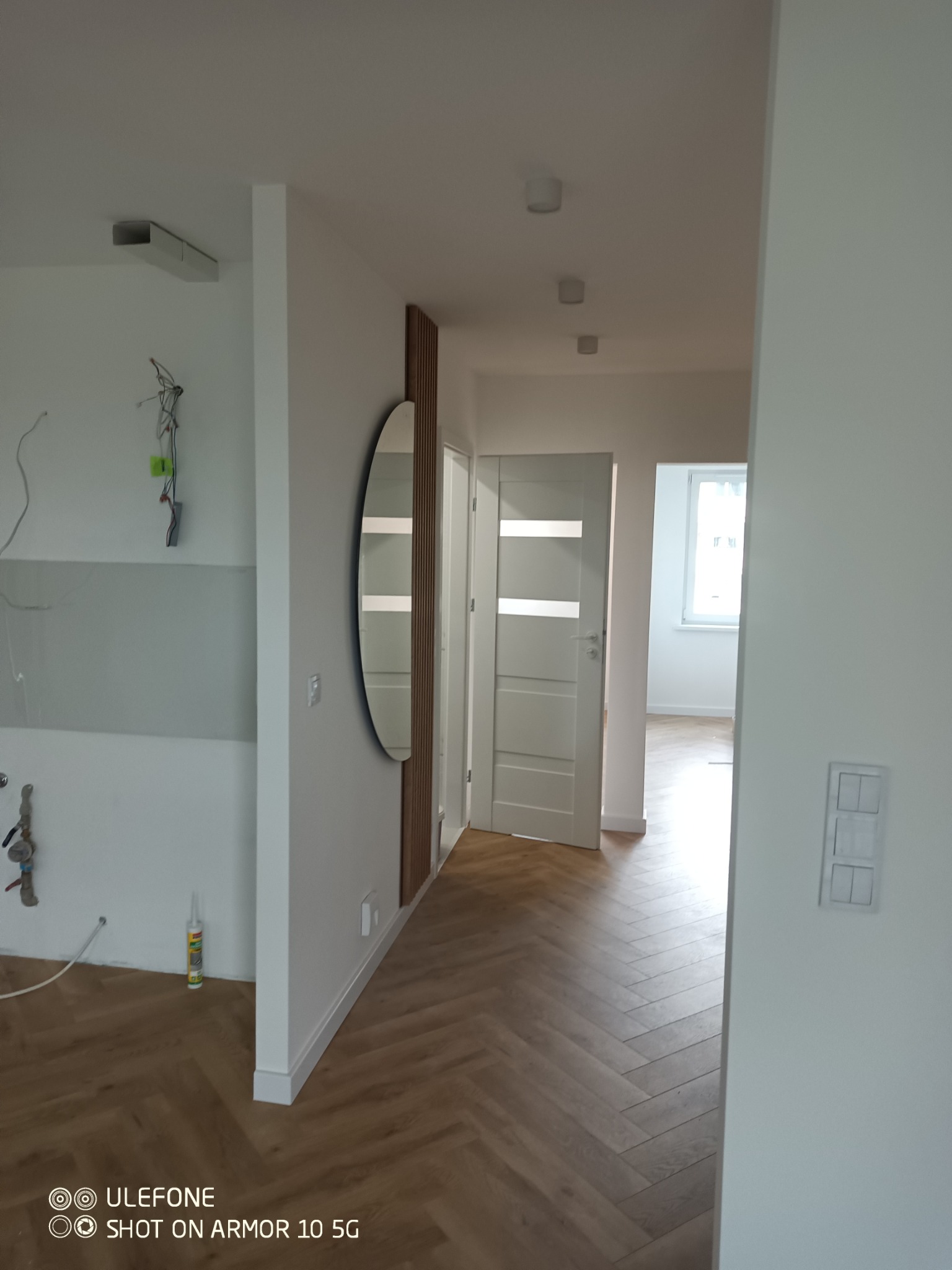 Nowoczesne wnętrze: parkiet w jodełkę, owalne lustro z drewnianą ramą, białe ściany i drzwi. Widoczne elementy instalacji elektrycznej w tle.