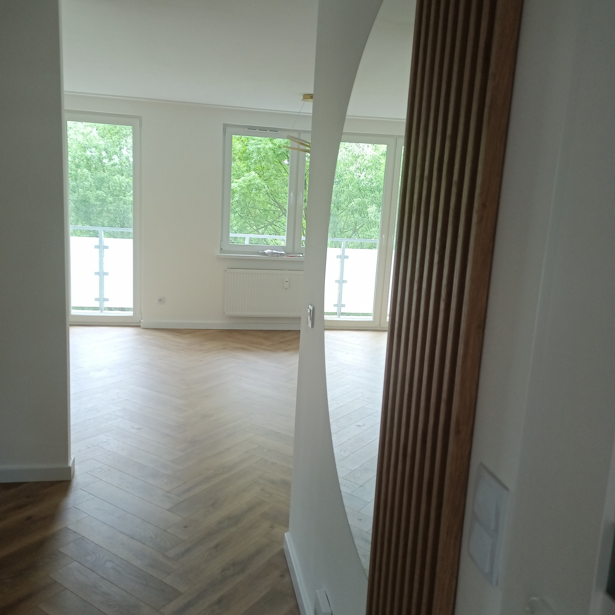 Jasne wnętrze z parkietem ułożonym w jodełkę, widoczne przez owalne lustro z drewnianą ramą. Okna z widokiem na zieleń. Minimalistyczny design.