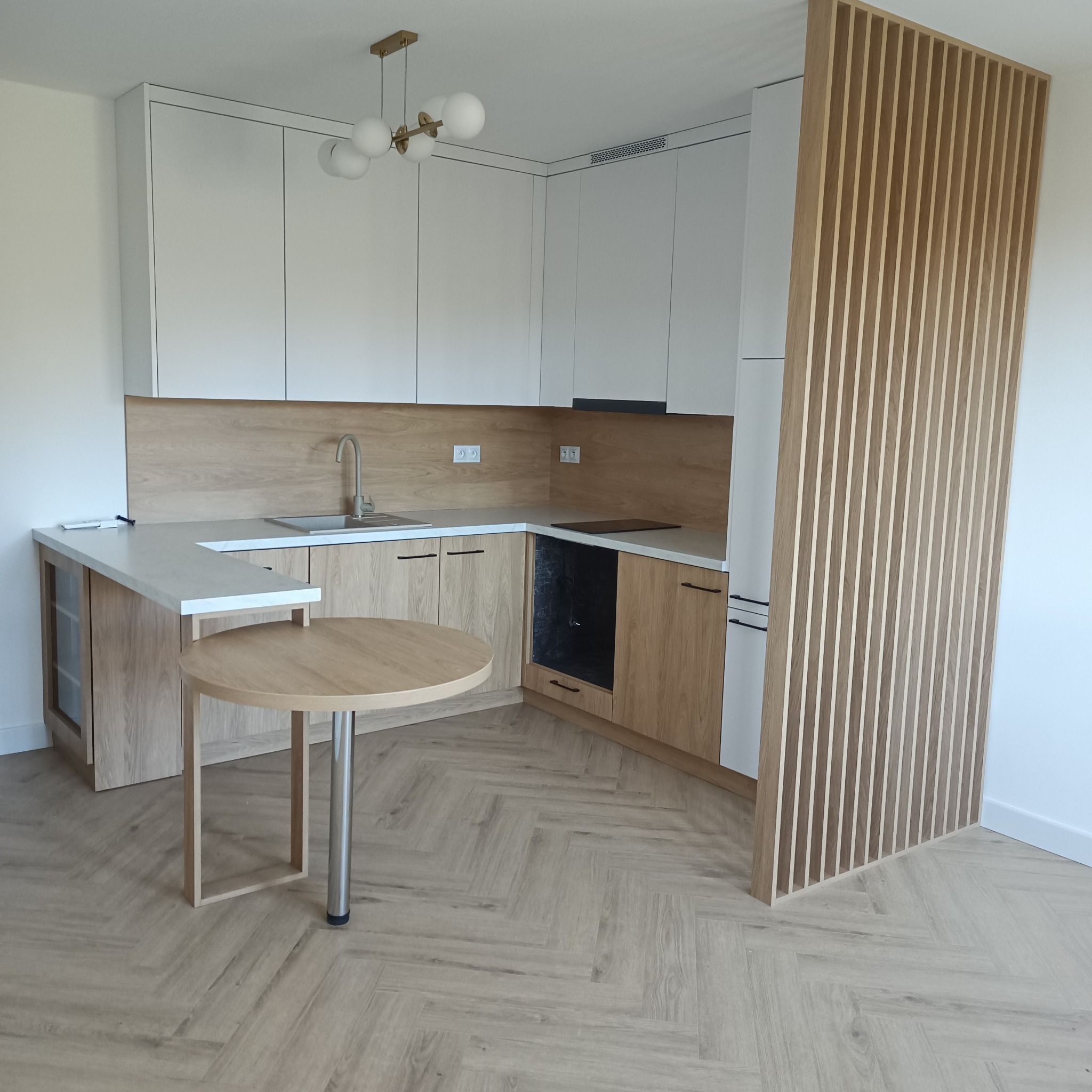 Jasna kuchnia z drewnianymi akcentami: szafki, blat i podłoga w jodełkę. Okrągły stół z metalową nogą. Dekoracyjna ścianka z drewnianych listewek.