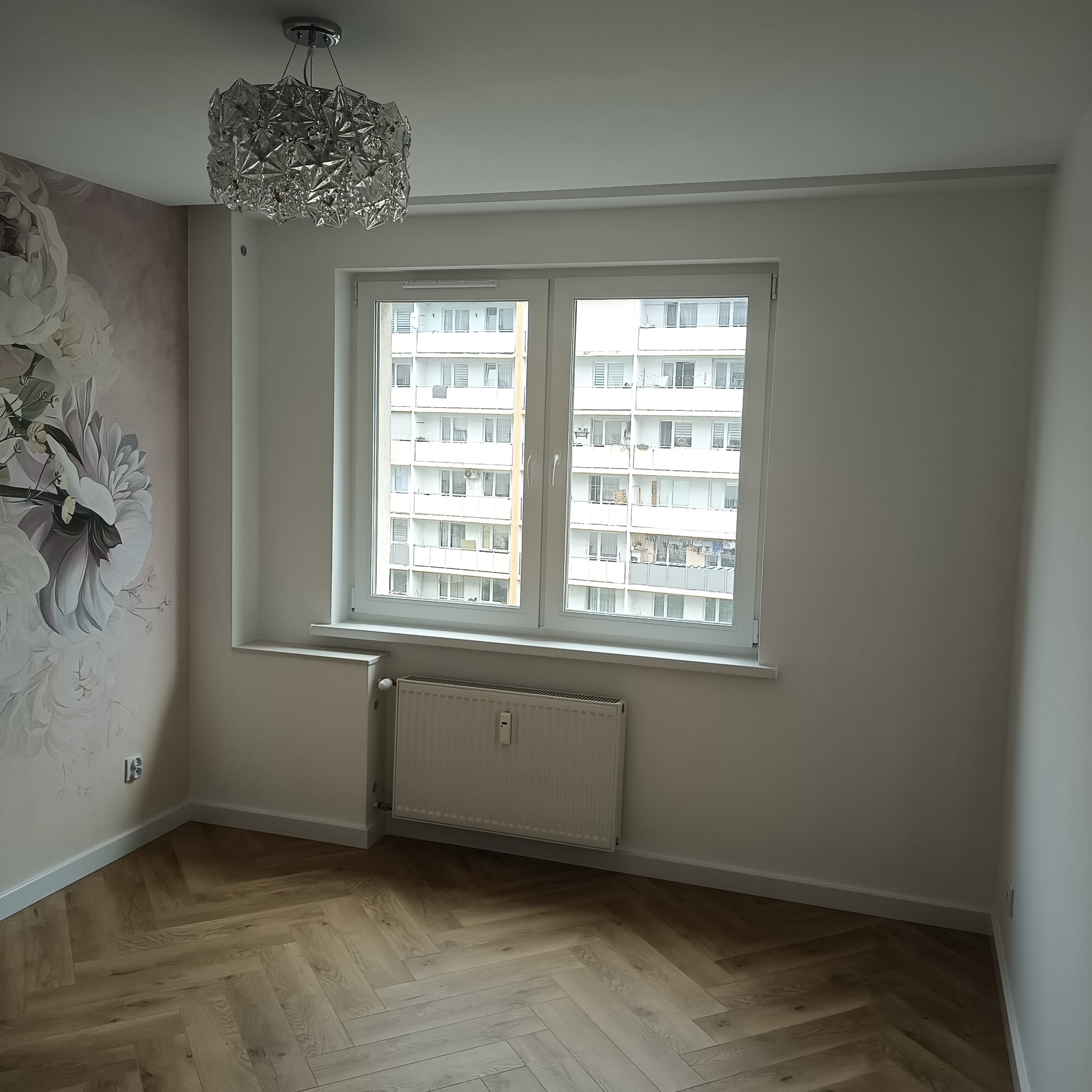 Jasne wnętrze po remoncie: parkiet w jodełkę, ściana z motywem kwiatowym, okno z widokiem na blok, nowoczesny żyrandol. Minimalistyczny design.