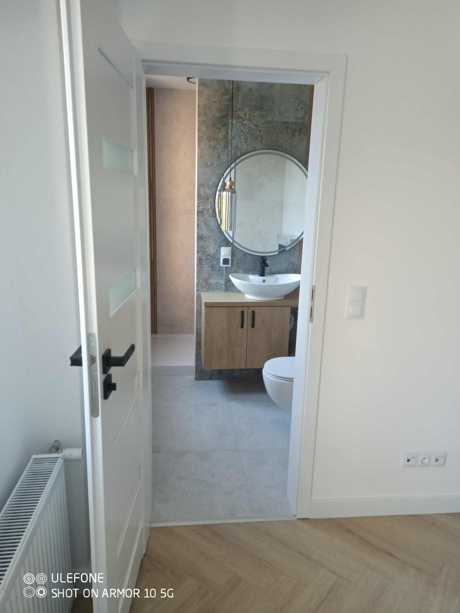 Nowoczesna łazienka z okrągłym lustrem nad umywalką na szafce, widoczna przez otwarte białe drzwi z czarną klamką. Minimalistyczny design, szare płytki, drewniane akcenty.