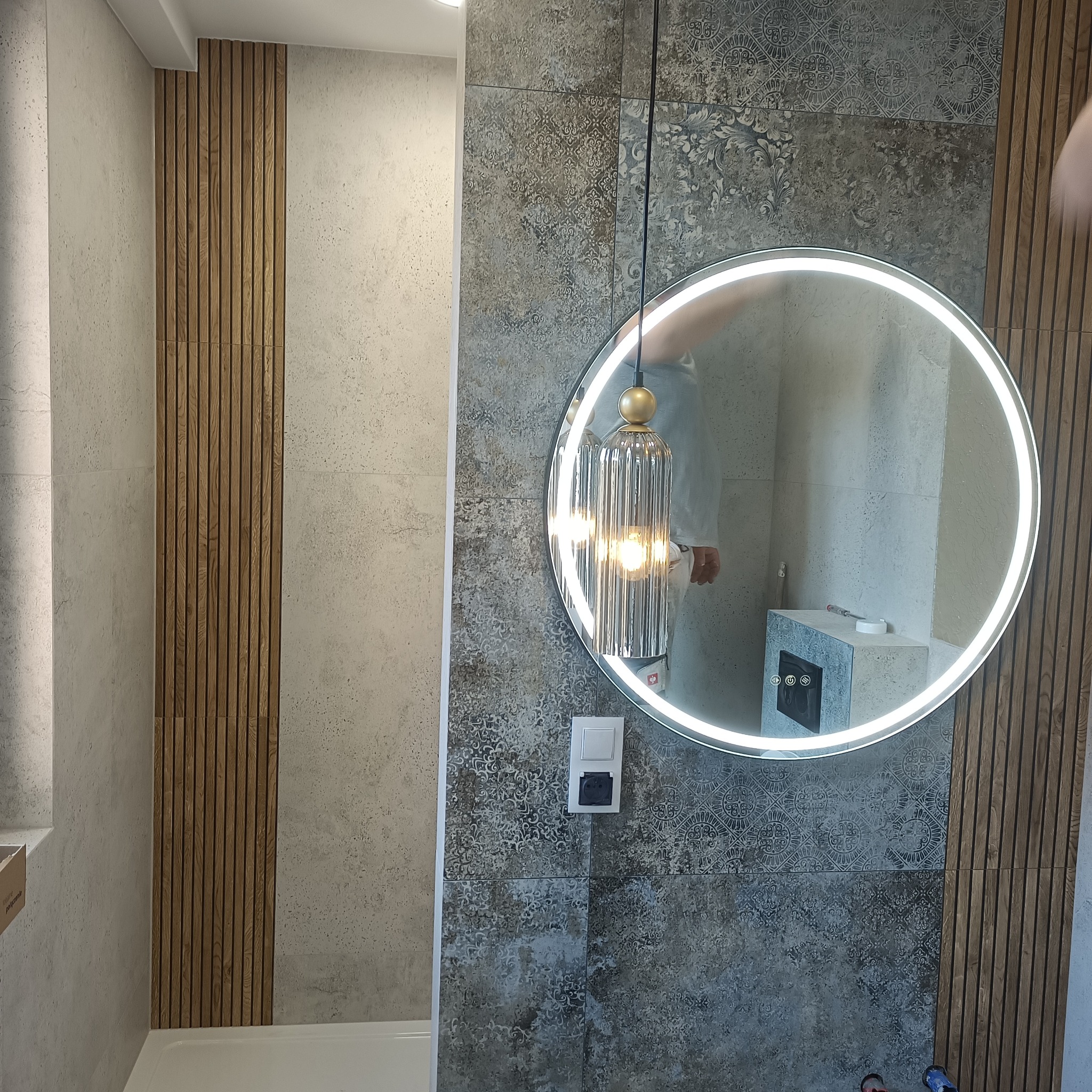 Wykończona łazienka z okrągłym lustrem LED, dekoracyjną lampą wiszącą i panelami ściennymi z drewna. Płytki w odcieniach szarości z wzorem.