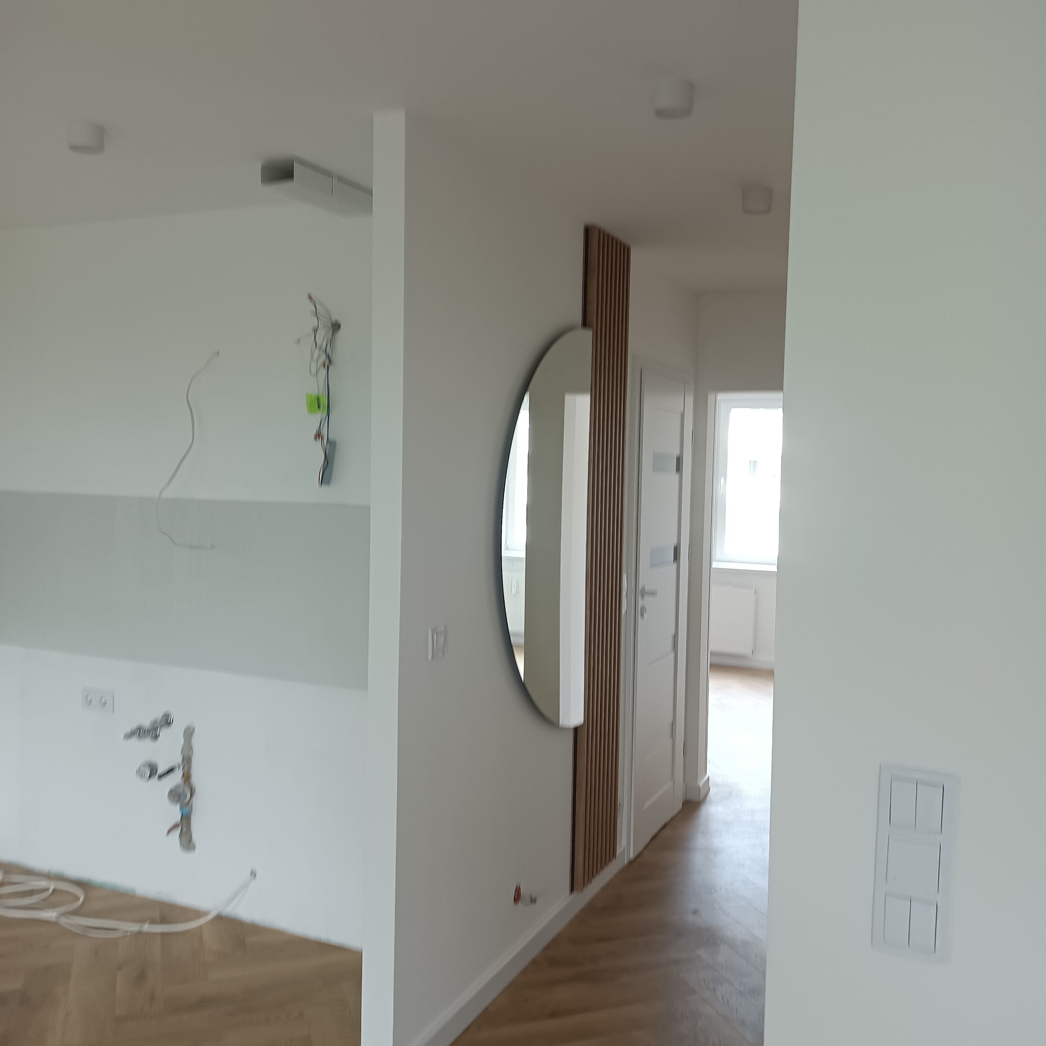 Wnętrze w trakcie wykańczania: owalne lustro z drewnianą dekoracją na ścianie, widoczne instalacje i podłoga. Minimalistyczny design w jasnych barwach.