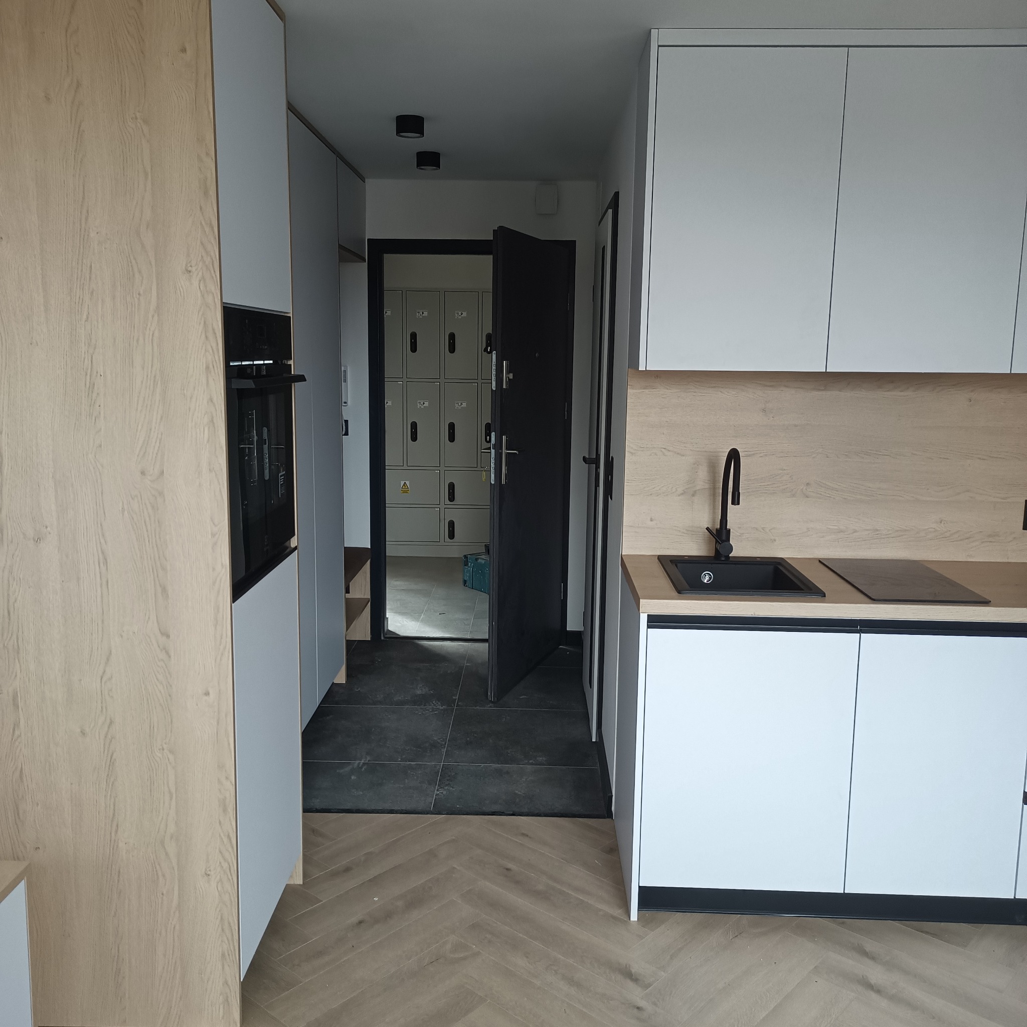 Nowoczesna kuchnia z białymi szafkami i drewnianym blatem, czarny zlew i bateria. Widok na korytarz z szafkami. Parkiet w jodełkę na podłodze.