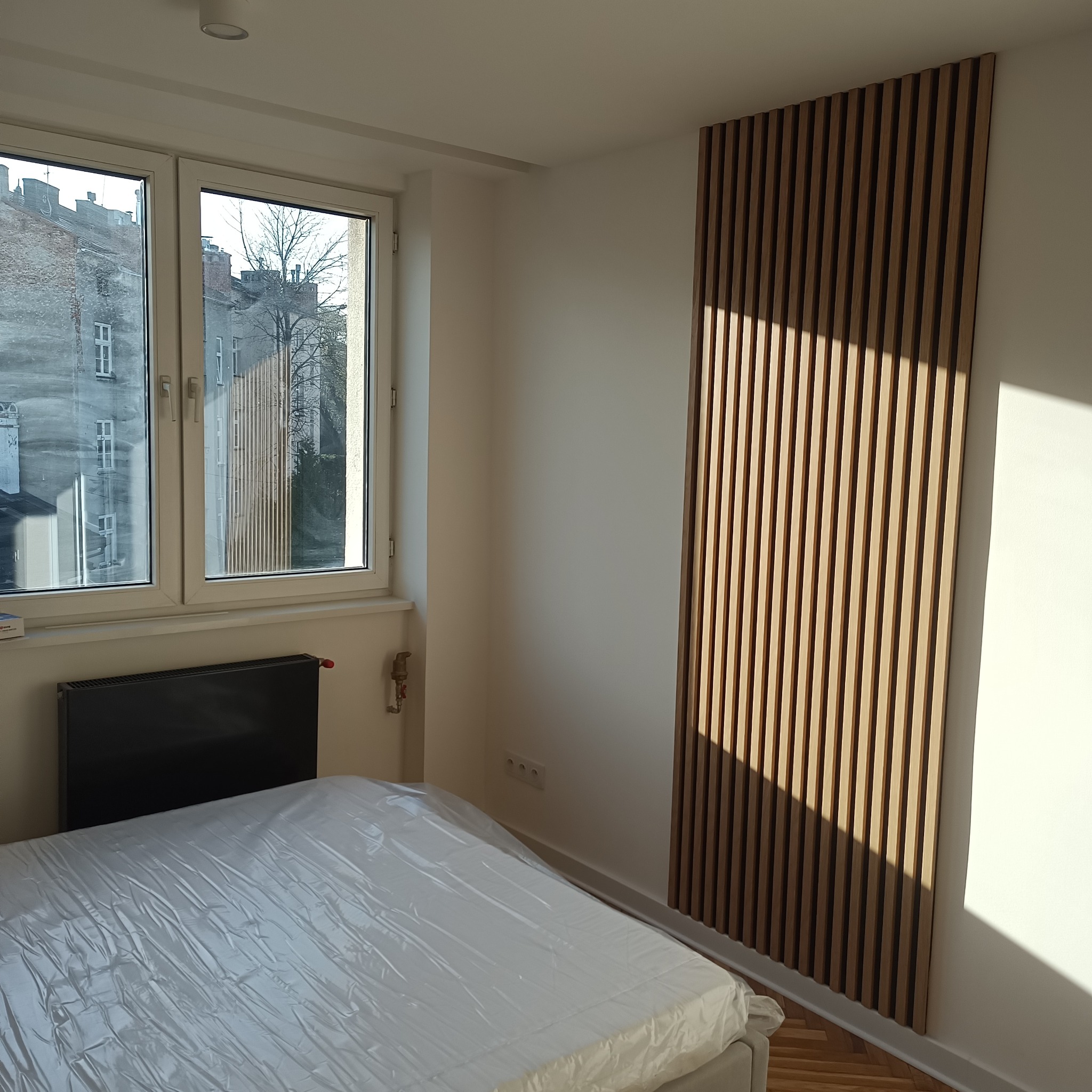 Nowoczesny pokój z drewnianą ścianką działową z listew, wpadające promienie słoneczne, widok z okna na kamienice. Minimalistyczny design wnętrza.