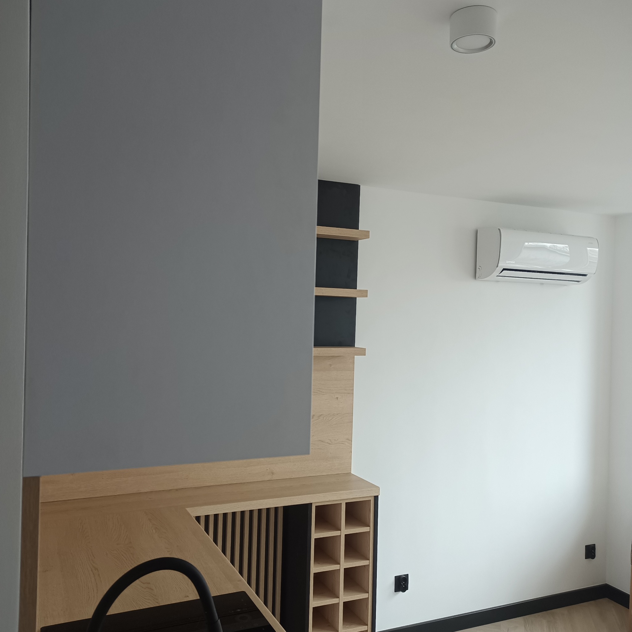 Nowoczesne wnętrze z szarą zabudową meblową, drewnianymi półkami i klimatyzatorem na białej ścianie. Minimalistyczny design z czarnymi akcentami.