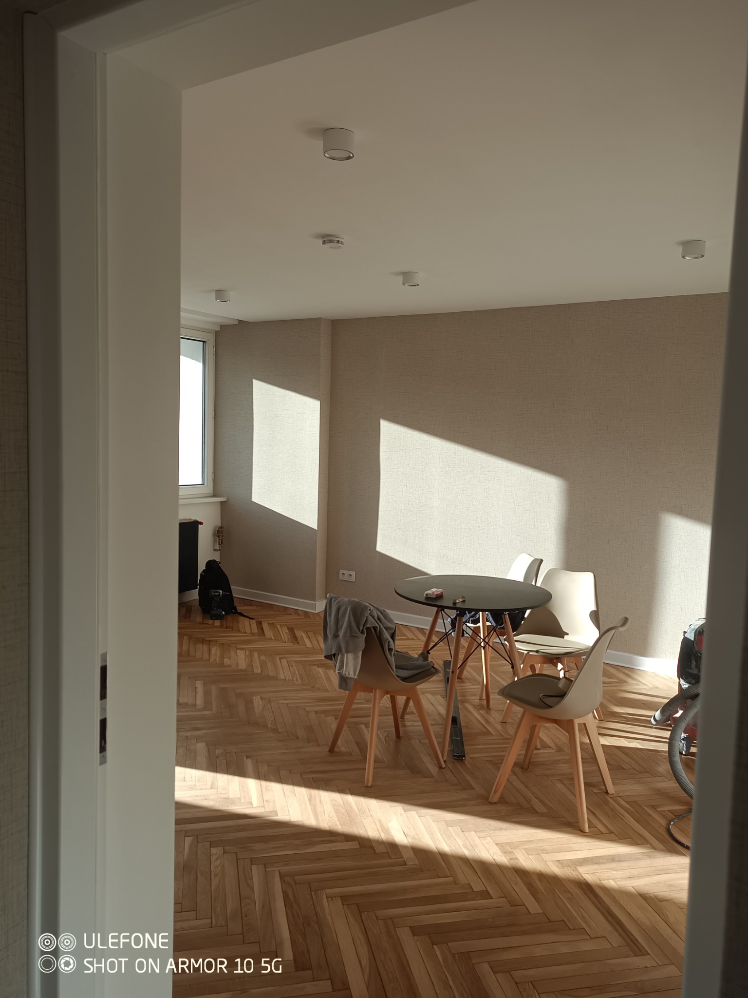 Jasne wnętrze z parkietem w jodełkę, widziane zza progu. Stół z krzesłami w świetle słonecznym. Minimalistyczny design, ciepłe odcienie, świeżo po remoncie.