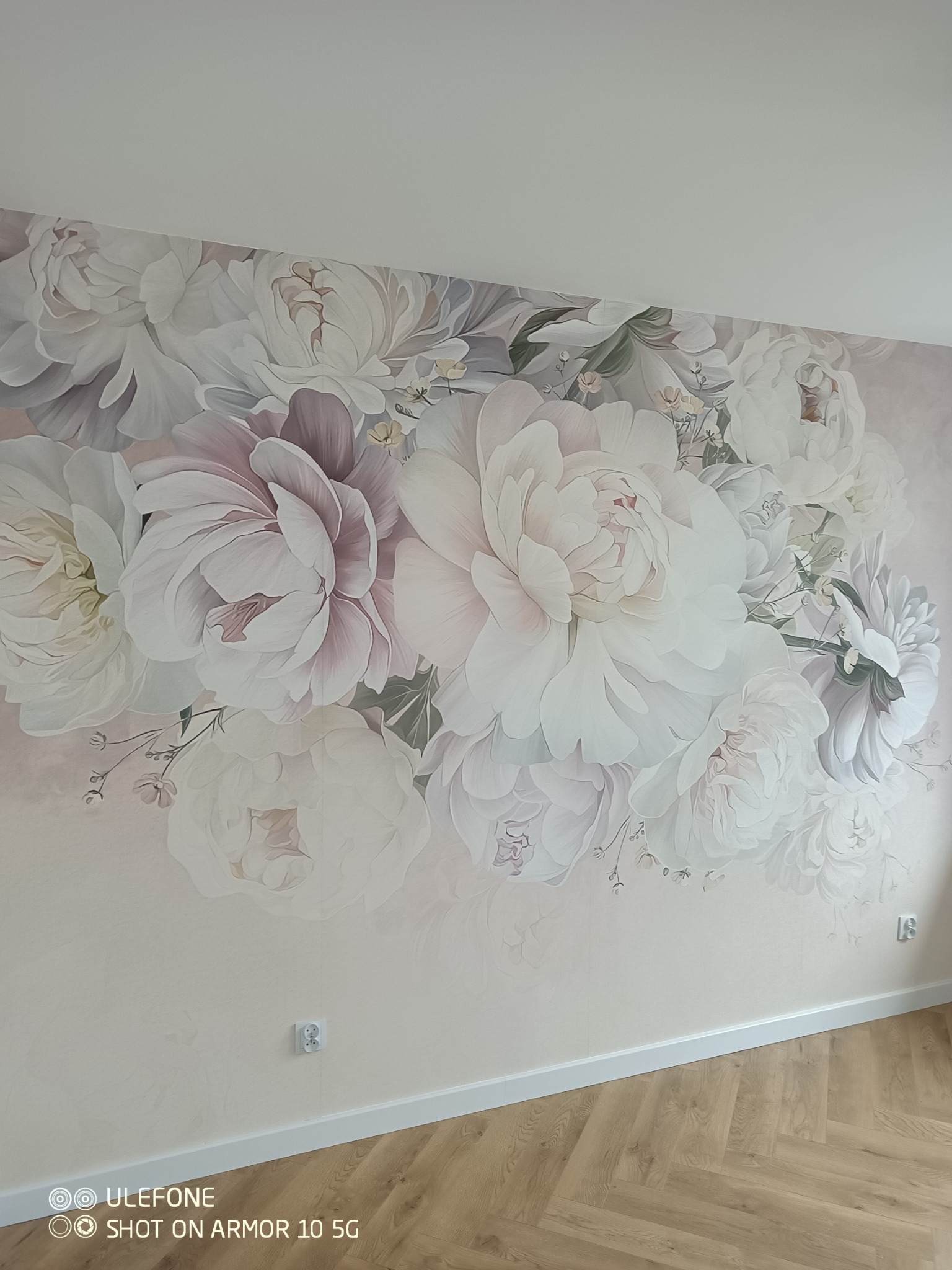 Ściana z fototapetą w kwiaty piwonii w pastelowych odcieniach, widoczny fragment podłogi z paneli w jodełkę. Dekoracja wnętrza w stylu romantycznym.