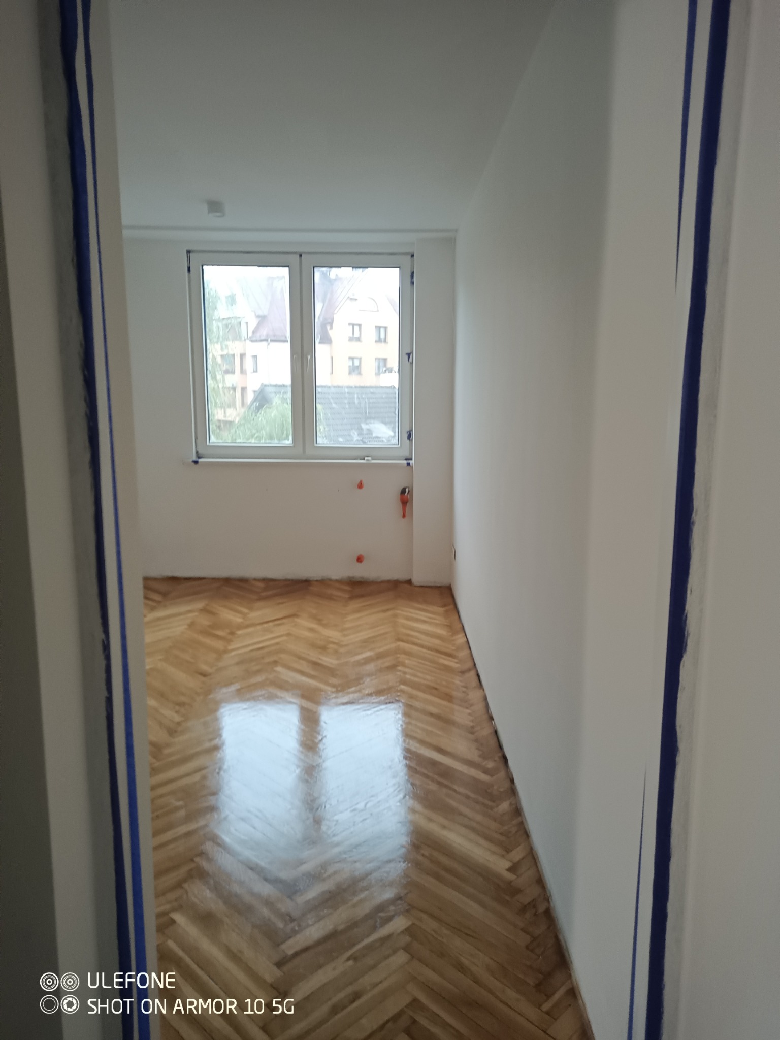 Pokój po remoncie: świeżo polakierowany parkiet w jodełkę, białe ściany z niebieską taśmą malarską przy framudze drzwi, okno z widokiem na budynki.
