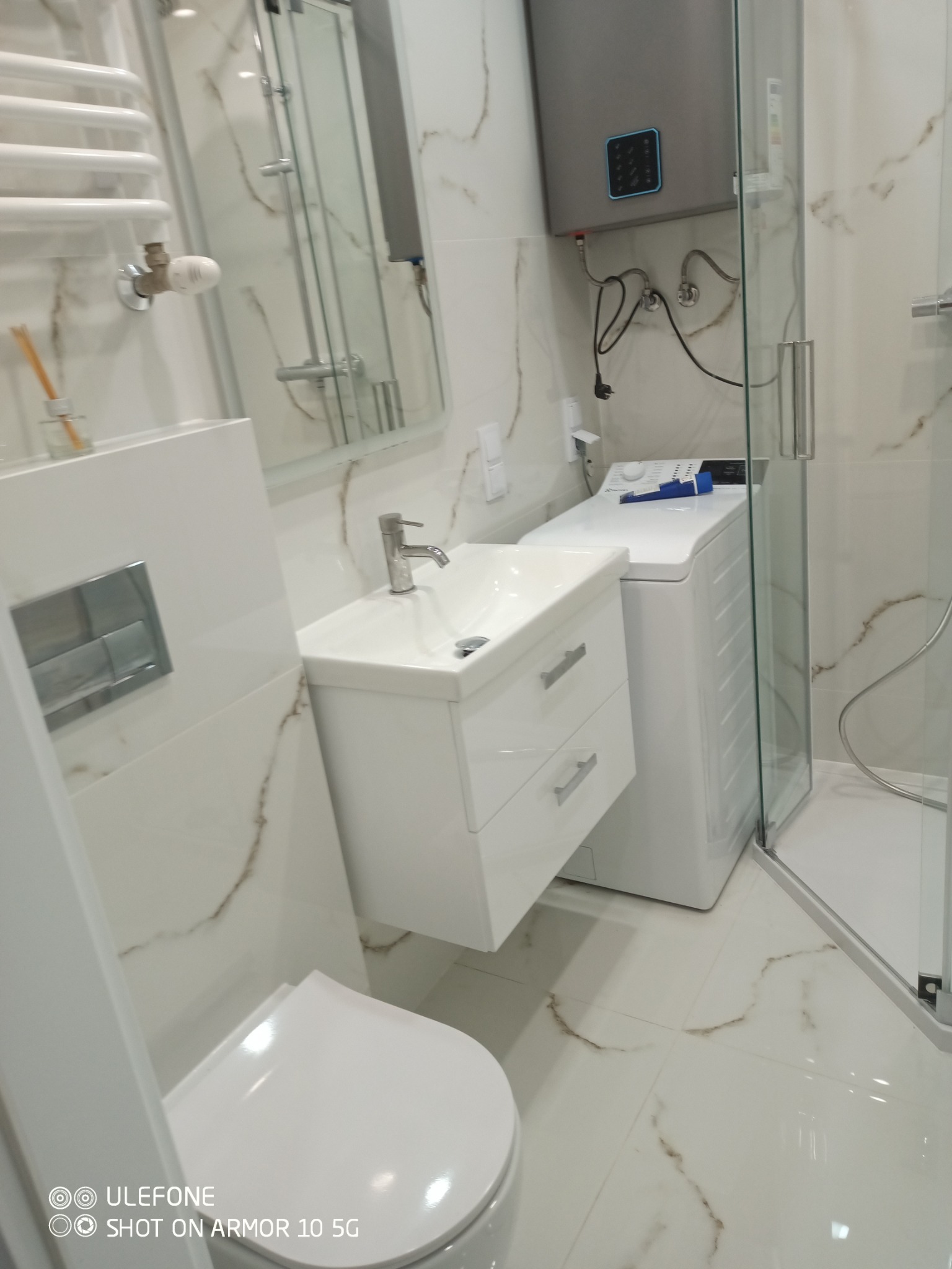 Biała, nowoczesna łazienka z umywalką na szafce, toaletą, pralką i kabiną prysznicową. Marmurowe płytki na ścianach i podłodze. Grzejnik drabinkowy.