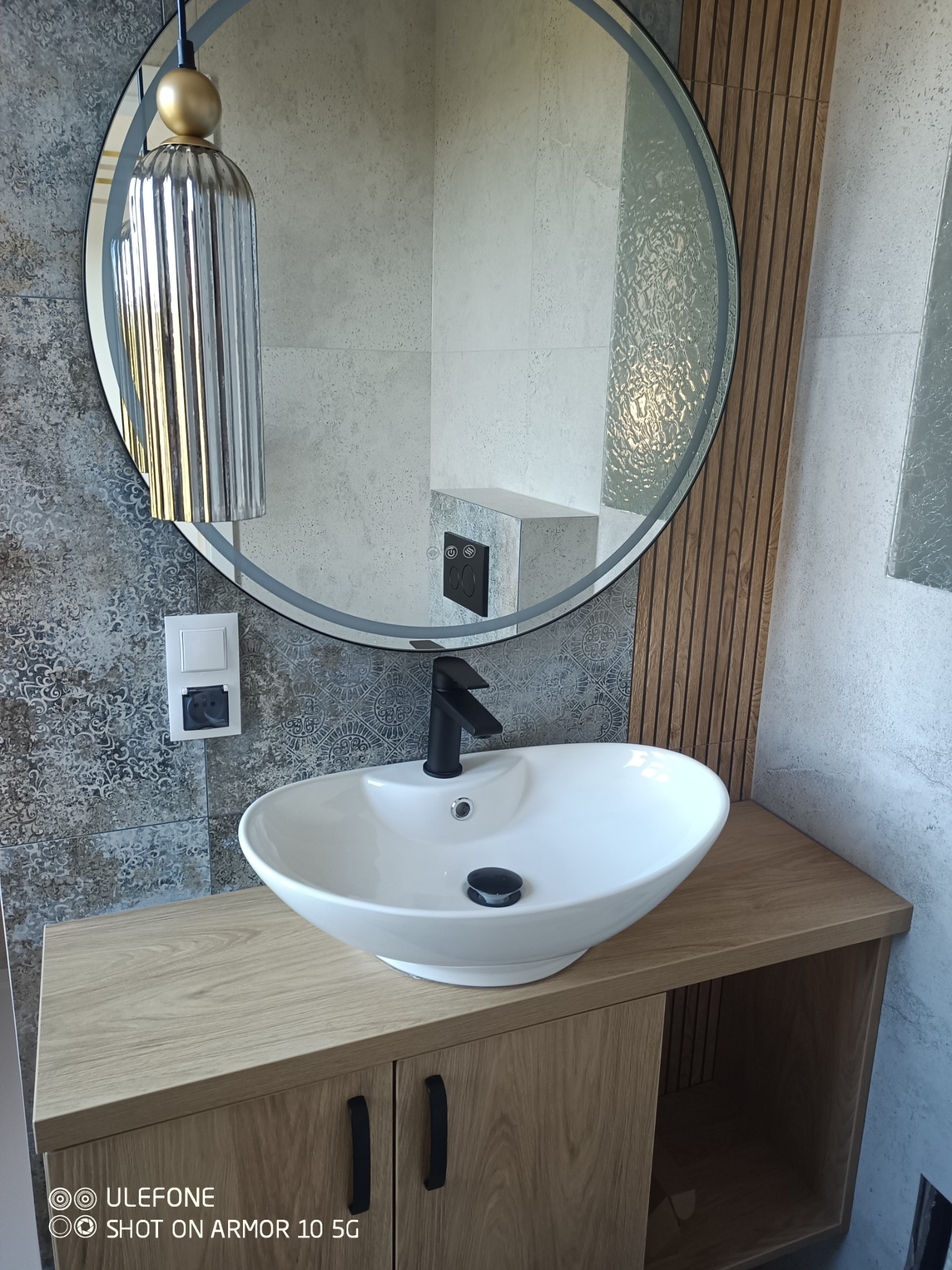 Nowoczesna łazienka z okrągłym lustrem, wiszącą lampą i umywalką nablatową na szafce z drewnopodobnym frontem. Na ścianie dekoracyjne panele i gniazdko elektryczne.
