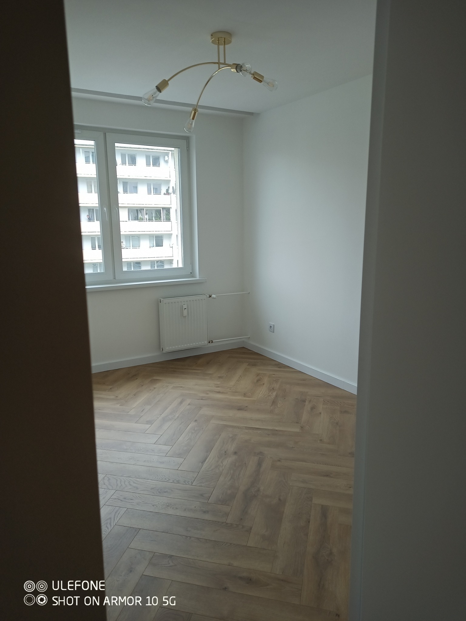 Pusty pokój z parkietem w jodełkę, białymi ścianami i oknem z widokiem na budynek. Nowoczesna lampa sufitowa. Minimalistyczne wykończenie wnętrza.