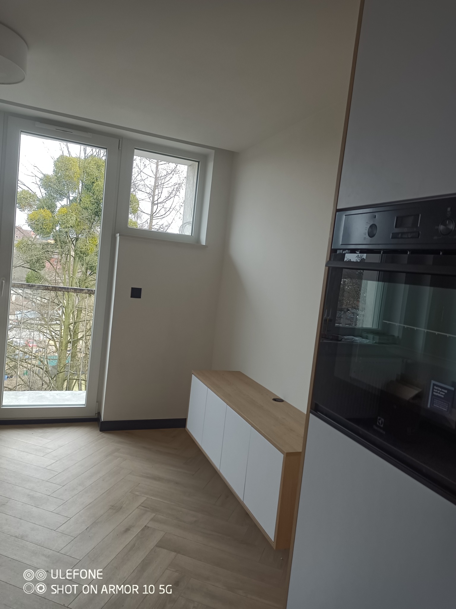 Jasne wnętrze z parkietem w jodełkę, zabudową kuchenną z piekarnikiem i szafką RTV pod oknem balkonowym z widokiem na drzewa z jemiołą.