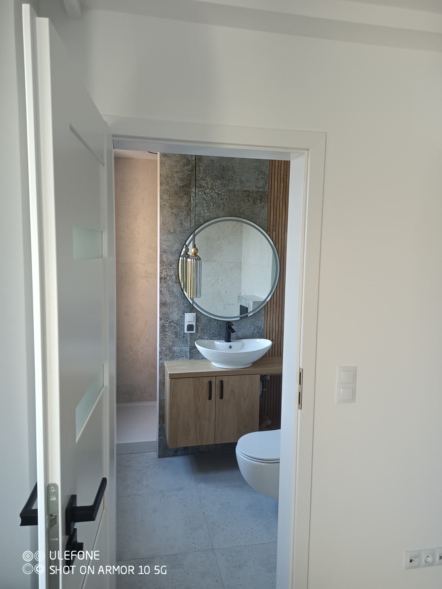 Nowoczesna łazienka z okrągłym lustrem, drewnianą szafką pod umywalką i szarą płytką na ścianie, widziana zza uchylonych białych drzwi.